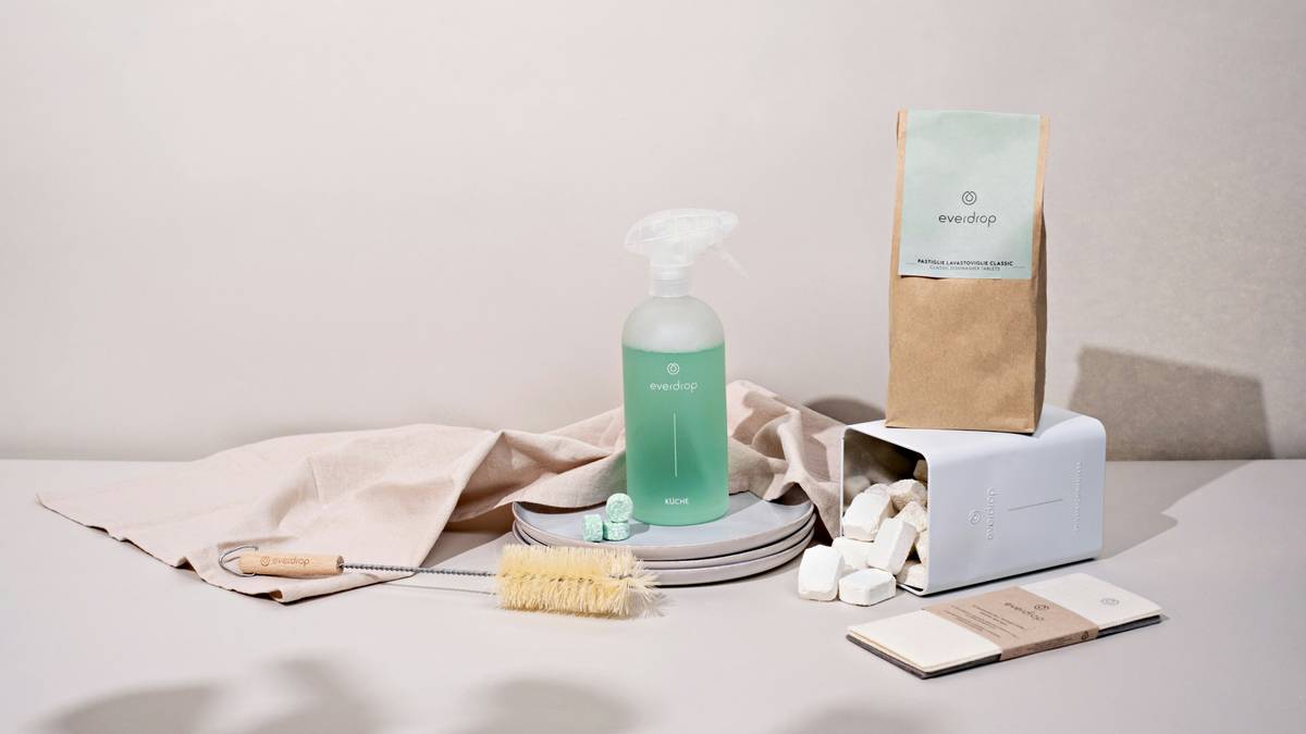 Hand holding a bottle of biodegradable dish soap next to other examples of biodegradable products like bamboo brushes and compostable sponges